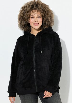 Kvinde med krøllet hår iført en sort fleece-hoodie med lynlås og mørke jeans, der smiler mod en ensfarvet hvid baggrund.