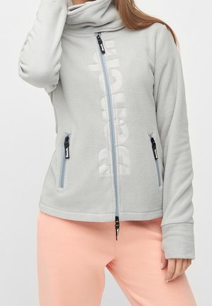 Woman wearing light grey zip-up fleece jacket with vertical logo and light pink pants, hand resting near face and waist.