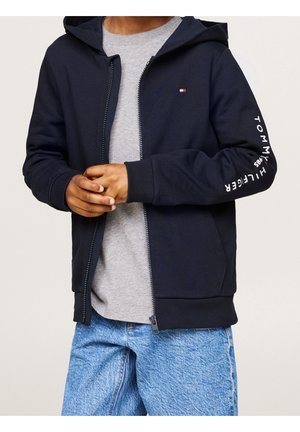 Osoba ubrana w rozpiętą granatową bluzę z małym logo na piersi oraz napisem "Tommy Hilfiger 1985" na rękawie, szarą koszulkę i niebieskie dżinsy.