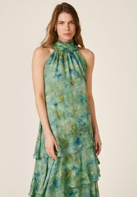 Femme portant une robe sans manches à col halter florale vert-bleu avec des couches à volants, debout sur un fond clair uni.