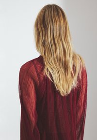 Durchsichtige burgunderfarbene Bluse mit vertikalen Falten, langen Ärmeln und fließender Silhouette, die aus einem strukturierten Material gefertigt ist und ein offenes Rücken-Design aufweist.