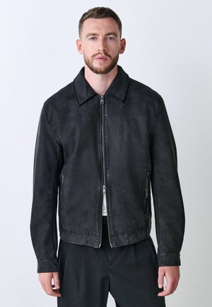 Man with short brown hair and beard wearing a black zip-up jacket and dark pants, standing against a plain light background.