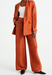 Rustově zbarvený lněný sako s uvolněným střihem a klopami, v kombinaci s kalhotami s širokými nohavicemi. Pod tím bílý žebrovaný top. Černé boty na jehlovém podpatku.