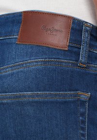 Tylnia kieszeń niebieskich dżinsów z denimu z brązową skórzaną etykietą marki przeszytą nad kieszenią oraz widoczną pętlą na pasek.