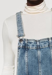 Farkkuhaalarit säädettävillä metallisilla soljilla. Kangas on vaaleansinistä pesua, jossa on haalistuneita kohtia ja sauman yksityiskohtia.