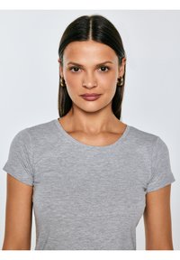 Kobieta o prostych ciemnych włosach, nosząca złote kolczyki koła i szarą koszulkę z krótkim rękawem, stoi przodem z neutralnym wyrazem twarzy.