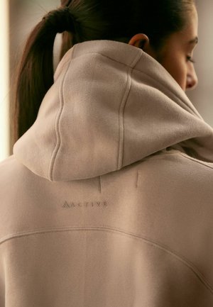 Beige hoodie met een zachte textuur, voorzien van een gestikt decolleté en het woord "ACTIVE" in reliëf op de bovenrug.