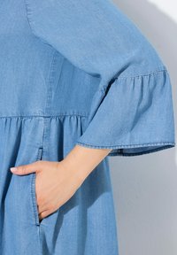 Světle modré denimové šaty s rozšířeným rukávem a volánkovým pasem. Obsahují boční kapsy a mají hladkou, jemnou texturu.