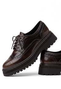 Paire de chaussures brogues en cuir marron foncé avec semelles épaisses à crampons noirs et fermetures à lacets sur fond blanc.