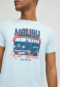 Világoskék pamut póló, amelyen egy vintage kisbusz grafikája látható, "MALIBU" szöveggel és "Ride Waves" szlogennel piros és sötétkék színben.
