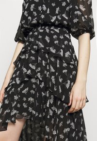 Schwarzes durchsichtige Kleid mit geschichteten Rüschen, weißem Blumenmuster und gepufften Ärmeln, gezeigt von der Taille bis zur Mitte des Oberschenkels auf der linken Seite einer Person.