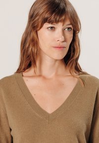 Pull en v khaki avec des bords côtelés, texture douce et coupe décontractée. Le tissu semble léger et légèrement duveteux.