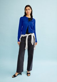 Blauer Cardigan aus Strickstoff, mit Knopfleiste vorne. Grauer Pullover, um die Taille gebunden, kombiniert mit hochgeschnittenen schwarzen Jeans und schwarzen Absatzsandalen.