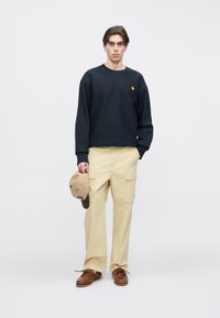 Σκούρο ναυτικό φούτερ με μικρό λογότυπο, μπεζ παντελόνι καμουφλάζ με πολλές τσέπες, καφέ παπούτσια τύπου boat shoes και καπέλο σε απόχρωση ταμπά. Απλός σχεδιασμός και χρώματα.