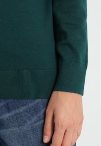 Vihreä neulottu paita, jossa on resorit hihansuissa ja helmassa, jonka päällä lepää käsi sinisellä farkkukankaalla. Pehmeä pinta ja istuva malli.