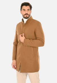 Cappotto di lana marrone con collo alto, tasche laterali e dettagli con bottoni, indossato sopra un dolcevita bianco, con una vestibilità su misura.