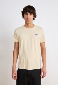 Beige pamut póló, kerek nyakkivágással és rövid ujjakkal. A bal mellen kék embléma található "ALPHA INDUSTRIES" felirattal.