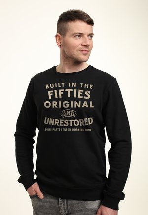 Mand iført sort sweatshirt med teksten "Bygget i halvtredserne, original og urestaureret, nogle dele stadig i funktion," hænderne i lommerne.