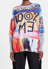 Färggrafisk oversized sweatshirt med en blandning av abstrakta mönster, djärva svarta bokstäver och kontrasterande gråa långärmar.