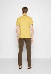 Žluté polo tričko s kontrastním lemováním, nošené s hnědými kalhotami a žabkami. Pohled zezadu, který ukazuje jednoduchý design a střih.