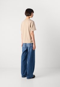 Persona in piedi di lato che indossa una camicia beige a maniche corte, jeans azzurri a gamba larga e scarpe nere su uno sfondo bianco.