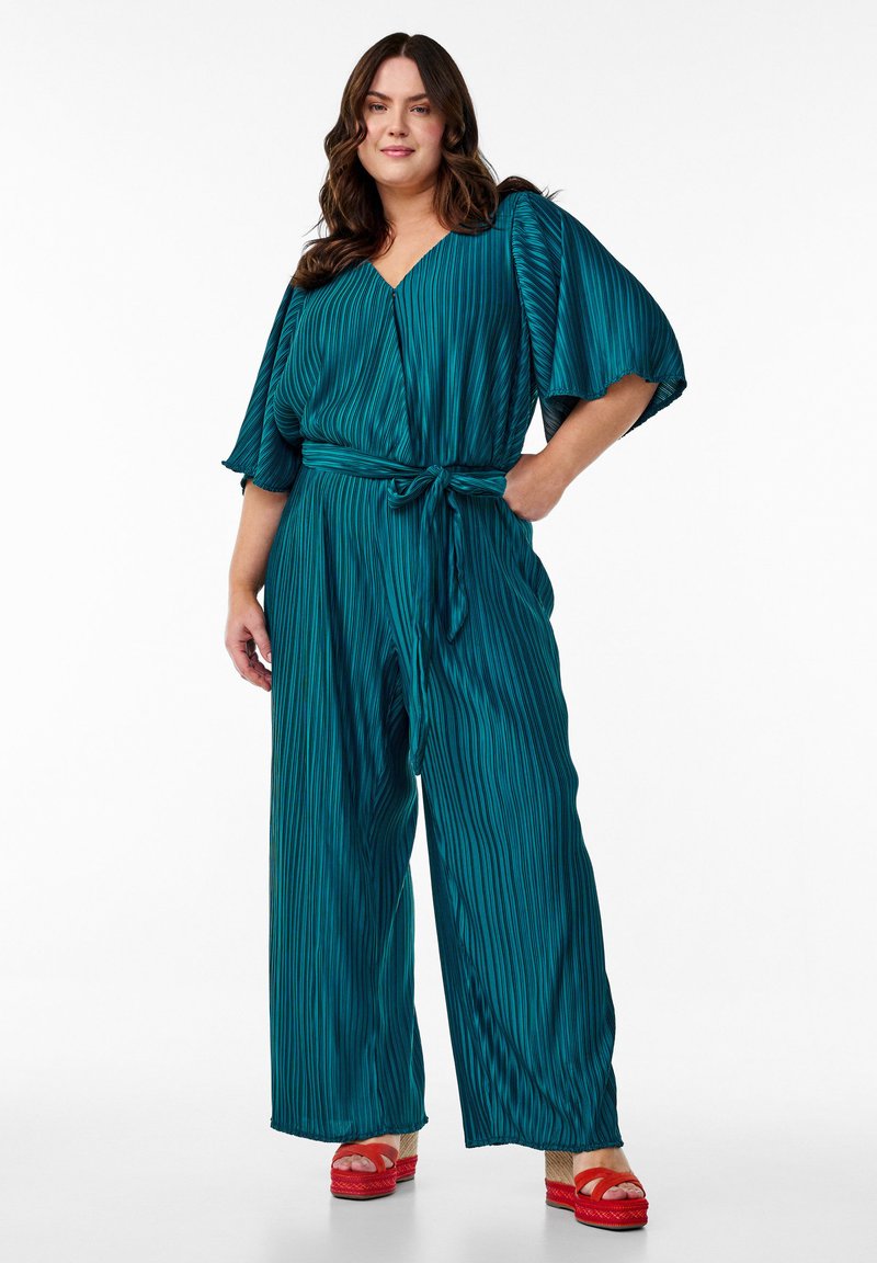 Türkisfarbener, plissierter Jumpsuit mit weiten Ärmeln und gebundenem Bund, kombiniert mit roten Plateausandalen. Verfügt über vertikale Streifen für zusätzliche Textur.