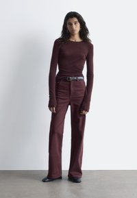 Burgunderfarbiges Longsleeve-Oberteil und hochgeschnittene, ausgestellte Hose, beide aus einem strukturierten Stoff; ein schwarzer Gürtel und Stiefeletten runden den Look ab.