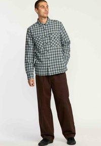 Camisa a cuadros en verde y blanco, de diseño abotonado con dos bolsillos frontales. Combinada con pantalones de pierna ancha marrones y zapatos negros.