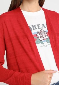 Rode gebreide cardigan met een gestructureerd patroon, open voorkant, gedragen over een wit grafisch T-shirt met een bloemenprint.