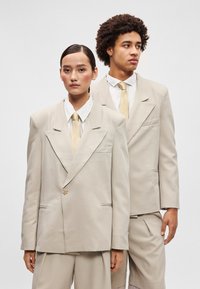 Fatos ajustados em bege claro com lapelas recortadas, fecho de um botão e acabamentos subtis; combinados com gravatas a condizer e camisas brancas.