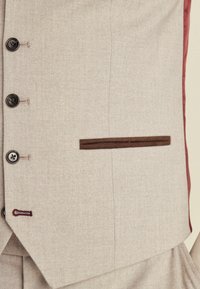 Beige Fischgrätenmuster-Weste mit dunklen Knöpfen, einem schmalen braunen Samt-Taschendetail und burdeosfarbigen Akzenten entlang des Innenfutters.
