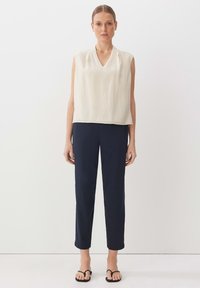 Top beige senza maniche con scollo a V, abbinato a pantaloni navy sartoriali che si stringono alle caviglie. Modella che indossa sandali neri piatti.