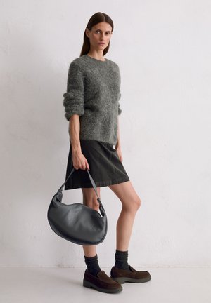 Frau in grauem, flauschigem Pullover, schwarzem Rock, dunklen Socken, Loafers, die eine große schwarze Ledertasche hält, vor einer weißen Wand.