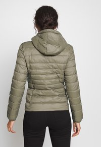 Oliwkowa kurtka ocieplana z odpinanym kapturem, pikowany wzór, długie rękawy i elastyczne mankiety, w zestawie z czarnymi spodniami.