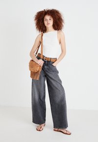 Balta topa bez piedurknēm, plata stila pelēki džinsi, brūns studēts jostas josta un gaiši brūns frēžas crossbody soma; modelis valkā brūnas siksniņu čības.