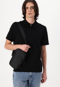 Svart polo-skjorte med glidelås i kragen, iført lyseblå jeans. Modellen holder en liten svart crossover-bag med glidelåsdetaljer.