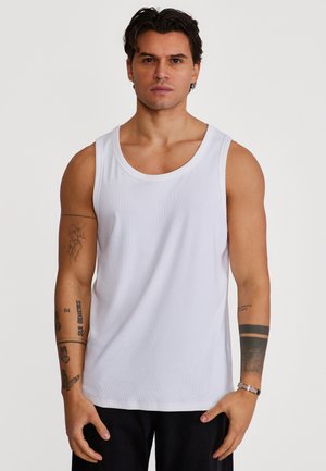 Mann med kort mørkt hår iført en hvit ermeløs skjorte, som viser forskjellige tatoveringer på begge armer, stående mot en ensfarget bakgrunn.