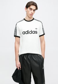 Mladý muž v bielom tričku Adidas s čiernymi pruhmi držiaci čiernu čiapku, nosiaci čierne nohavice, stojí pred jednoduchým svetlým pozadím.