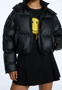 Veste matelassée noire avec une fermeture éclair à l'avant, un col montant et un détail en velcro, portée sur un t-shirt graphique. Texture lisse et brillante.