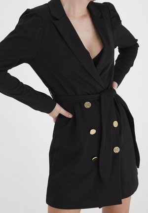 Frau trägt ein schwarzes, tailliertes Blazer-Kleid mit goldenen Knöpfen, die Hände in die Hüften gestützt, und zeigt einen tiefen V-Ausschnitt auf einem einfarbigen hellen Hintergrund.
