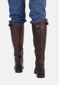 Bottes en cuir marron hautes avec une texture lisse, des zippers sur les côtés et des boucles décoratives en haut, associées à un jean en denim bleu.