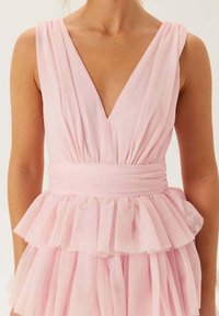 Jasnoróżowa sukienka bez rękawów z głębokim dekoltem w kształcie litery V, marszczonym gorsetem i wielowarstwową falbaną na spódnicy. Miękki, lejący się materiał o gładkiej fakturze.