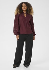 Blouse bordeaux avec des manches bouffantes en dentelle, en tissu léger et avec un petit col en V. Associée à un pantalon noir ample et des chaussures plates noires.