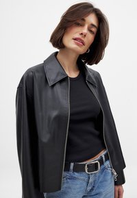 Juodas odinis švarkas su plačiu apykakle, sidabrinės užtrauktuko detalės, dėvimas ant juodo įgulo viršaus ir mėlynų džinsų, demonstruojantis neoficialų stilių ir elegantišką dizainą.