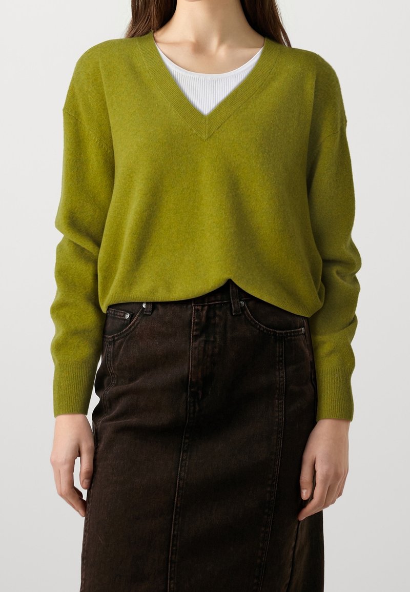 Mujer vestida con un suéter verde oliva de cuello en V sobre una blusa blanca, combinado con una falda de mezclilla negra de cintura alta.