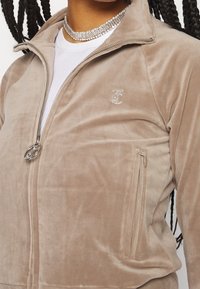 Beige zipjacka tillverkad av mjukt, plyschmaterial; har en subtil logotyp, sidoficka och en slät textur med silverfärgade detaljer.