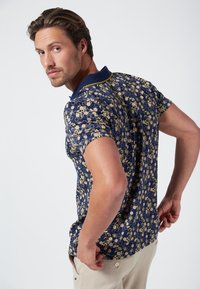 Marineblauwe poloshirt met een geometrisch patroon in goud en room. Beschikt over een effen marineblauwe kraag met gele rand. Korte mouwen en een slim fit ontwerp.