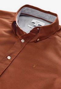 Chemise boutonnée couleur rouille avec boutons blancs, col intérieur rayé et petit logo brodé de cerf sur la poitrine.