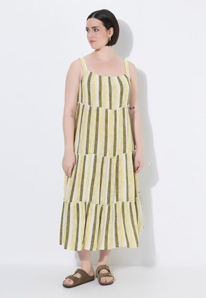 Frau mit kurzen dunklen Haaren, die ein ärmelloses Kleid mit gelben, weißen und olivgrünen vertikalen Streifen und braune Sandalen mit Schnalle trägt, steht vor einem weißen Hintergrund.