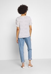 Mujer con cabello rizado, vestida con una blusa blanca de lunares de manga corta, jeans claros arremangados y sandalias de tacón blancas, de pie con la espalda hacia la cámara.